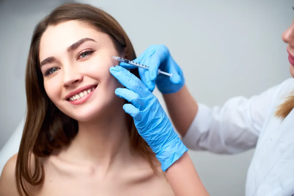 Blue-gloved hands position a syringe near a smiling woman’s outer cheek.