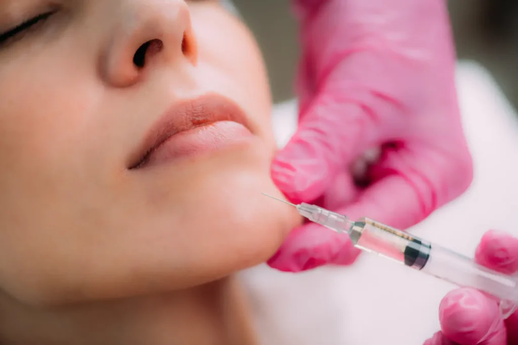 Pink-gloved hands hold a syringe with the needle positioned near a woman’s chin.